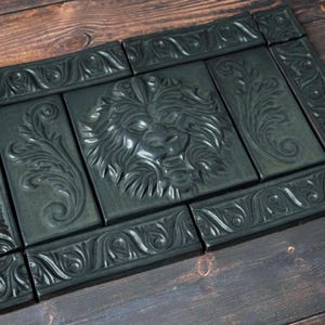 May include: A dark green, rectangular decorative tile set with a lion's head and floral relief carvings. The tiles are arranged to form a larger rectangular shape, with ornate borders. The set is displayed on a dark wood surface.