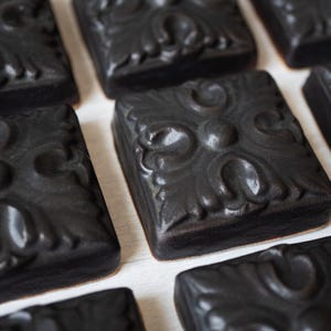 May include: A collection of dark grey, square soaps with embossed floral patterns. The soaps are arranged in a grid, highlighting their intricate designs and textures. The soaps are a uniform colour, and the image is a close-up.