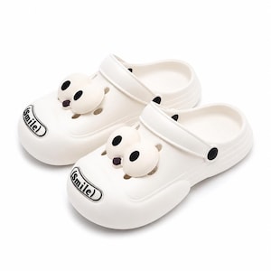 May include: A pair of off-white slip-on clogs featuring a rounded toe, a pivoting heel strap, and two large, cartoon-style eyes attached to the top. The front of each clog displays a black and white rectangular badge with the word Smile.