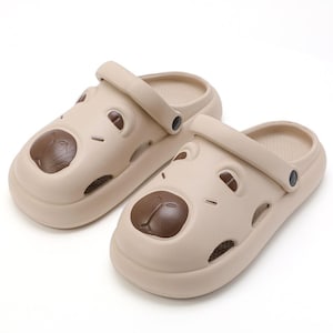 May include: A pair of beige slip-on clogs featuring a molded bear face design on the front. The shoes have a rounded toe, a pivoting heel strap with dark circular accents, and cut-out ventilation holes along the sides for breathability.