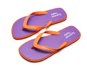 May include: A pair of purple flip-flops with orange straps. The footbed features a textured surface and the white text Summer Taste printed on the heel area and on one of the orange straps. The soles are orange.