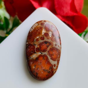 May include: An oval gemstone featuring a mix of reddish-brown, tan, and black patterns. The polished surface highlights the natural colour variations. This stone is likely intended for jewellery or decorative use. The gemstone measures approximately 3 cm in length.