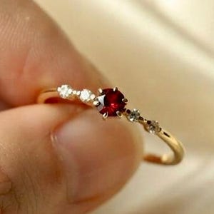 May include: A thin gold ring featuring a central round-cut deep red gemstone flanked by two smaller clear stones on each side. The band is slender and polished, showcasing a delicate design with a prong setting for the primary red stone.