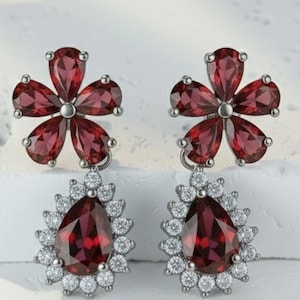 Peut inclure: Boucles d'oreilles en argent avec un motif floral et des pierres précieuses rouge foncé. Chaque boucle d'oreille a une partie supérieure en forme de fleur et une partie inférieure en forme de goutte d'eau, entourée de petites pierres claires. Présentées sur une surface blanche.