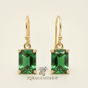 Puede incluir: Un par de pendientes colgantes dorados con piedras preciosas verdes esmeralda. Los pendientes tienen un diseño simple y elegante con cierre de gancho. El texto "IQRGEMSSHOP" es visible en la parte inferior.