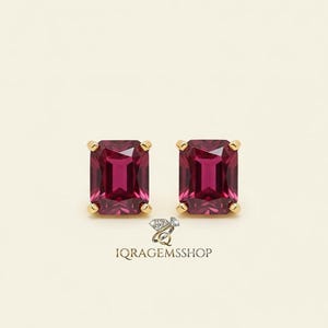 May include: A pair of gold-colored stud earrings, each featuring a rectangular, deep red gemstone. The earrings are set against a cream-colored background. The text "IQ RAGEMS SHOP" is visible at the bottom.