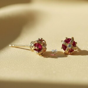 Op de afbeelding: Een paar goudkleurige oorstekers met een kroonontwerp. Elke oorbel heeft een ronde, dieprode edelsteen in het midden, omringd door gouden filigraan en kleine heldere accentstenen. De oorbellen zijn geplaatst tegen een zachte, neutrale achtergrond.