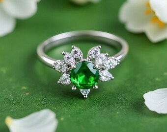 Puede incluir: Un anillo plateado con una piedra preciosa verde vibrante en el centro, rodeada de piedras claras más pequeñas. El diseño del anillo se asemeja a una flor. El fondo es verde suave con pétalos de flores blancas.