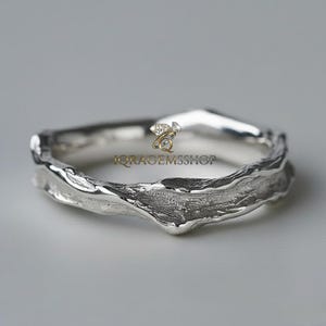 May include: A silver ring with a textured, organic design. The ring has a unique, irregular shape, with a rough, bark-like texture. The ring is set against a neutral background, highlighting its intricate details. The ring is a piece of jewellery.