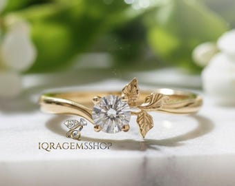 May include: A gold ring with a round, clear gemstone and leaf accents. The ring features a delicate design with a band that curves around the finger. The ring is set against a blurred background of green foliage and white flowers. The text "IQRAGEMSSHOP" is visible.