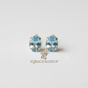 May include: A pair of oval-cut, light blue gemstone stud earrings set in gold. The earrings are displayed on a white background. The logo "IQRA GEMSSHOP" is at the bottom.