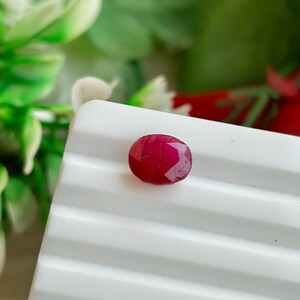 May include: A faceted, oval-shaped ruby gemstone. The ruby is a deep, rich red color and sits on a white, ridged surface. The background features blurred green and red foliage.