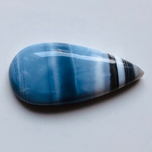 Può includere: Una gemma lucida a forma di goccia con un colore di base azzurro chiaro. La pietra presenta bande bianche, nere e grigie, creando un motivo a strisce. La gemma è liscia e riflettente, con una finitura lucida. La forma generale è allungata e simmetrica.