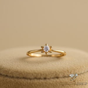 May include: A delicate gold ring featuring a six-petal flower design with a central, sparkling diamond. The ring's band is slender, and the setting is a bright, polished gold. The ring is displayed on a neutral, textured surface.