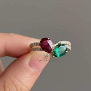 May include: A gold ring with a pear-shaped ruby and emerald gemstone design. The ring has a double band with small, clear accent stones. The gemstones are set close together, creating a unique and elegant look. The ring is held by a person.