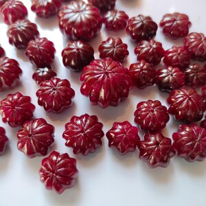 Può includere: Una collezione di piccole perline a forma di fiore, di colore rosso scuro. Le perline hanno un design sfaccettato, che cattura la luce e crea un effetto scintillante. Sono sparse su una superficie bianca, suggerendo un tema artigianale o di gioielleria.