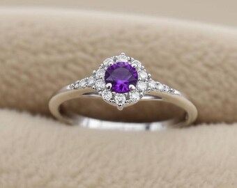 May include: A silver ring featuring a round, deep purple gemstone surrounded by a halo of smaller, clear stones. The ring's band is slender and polished, with additional small stones set on either side of the central design.