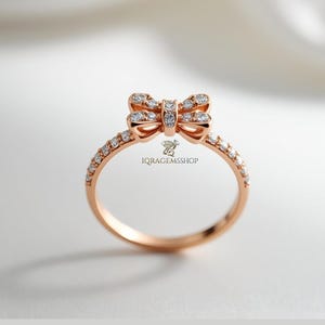 Puede incluir: Anillo de oro rosa con un diseño de lazo adornado con pequeñas y brillantes piedras preciosas. La banda también está adornada con diminutas piedras preciosas. El anillo se presenta sobre un fondo suave y neutro, resaltando sus delicados detalles. El anillo es de IQRAGEMSSHOP.