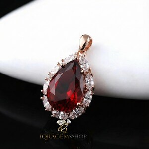 May include: A rose gold pendant with a large, pear-shaped, deep red gemstone at its center. The gemstone is surrounded by smaller, clear, oval-cut stones. The pendant has a loop at the top for a chain. The bottom of the pendant has the text "IQ RAGEMSSHOP".