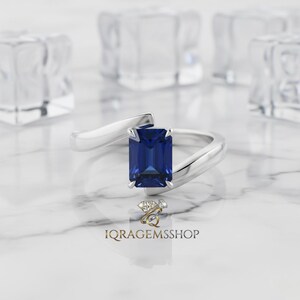 May include: A silver ring featuring a large, rectangular, deep blue gemstone. The ring's band has a curved design, creating a modern, elegant look. The ring is set against a blurred background with ice-like shapes. The shop name "IQRA GEMSSSHOP" is visible.