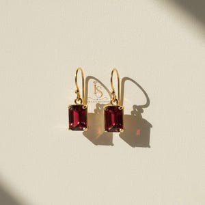 Puede incluir: Un par de pendientes colgantes dorados con piedras preciosas rectangulares de color rojo intenso. Los pendientes se muestran sobre un fondo claro, con una sombra a la derecha. Diseño sencillo y elegante.