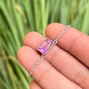 Pode incluir: Uma pulseira de prata com uma pedra preciosa rosa vibrante retangular. A pedra é colocada em uma moldura de prata e conectada a uma corrente delicada. A pulseira é exibida em um fundo verde desfocado.