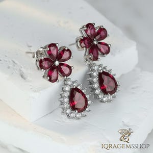 Puede incluir: Pendientes de plata con diseño floral y pétalos rojo oscuro. Cada pendiente tiene una gema roja en forma de lágrima rodeada de pequeñas piedras transparentes. Los pendientes están sobre un fondo blanco.