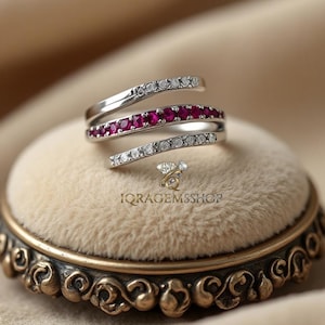 Può includere: Un anello argentato con un design a spirale. L'anello presenta una fila di piccole gemme rosse rotonde e una fila di gemme chiare rotonde. È esposto su una superficie di velluto beige. Il testo "IQGEMS SHOP" è visibile.