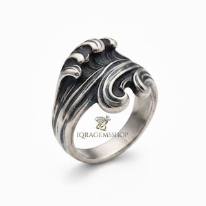 May include: A silver ring featuring a detailed wave design. The ring's wave crests are highlighted with darker shading, creating depth. The band is smooth and polished, with the text "IQ RAGEMSSHOP" visible on the inside.