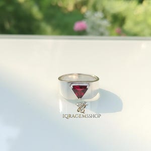 May include: A silver ring with a triangular, deep red gemstone. The ring has a wide band and a minimalist design. The gemstone is set in a bezel setting. The ring is displayed on a white surface with a blurred green background.