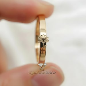 May include: A gold-colored ring featuring a detailed lotus flower design. The ring is held up, showcasing the delicate floral engraving. The ring's band is smooth and polished, reflecting light. The shop's name, IQRAGEMSSHOP, is visible at the bottom.