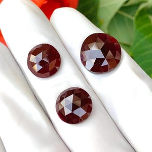 Può includere: Tre gemme rotonde, rosso scuro, sfaccettate. Le gemme sono esposte su una superficie bianca, con un fiore rosso e fogliame verde sullo sfondo. Le gemme hanno un taglio a rosa, con molteplici sfaccettature che riflettono la luce.