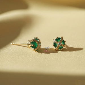 Puede incluir: Un par de pendientes dorados con piedras preciosas de color verde esmeralda. Los pendientes tienen un diseño en forma de corona con pequeñas piedras de acento transparentes. Las piedras preciosas miden aproximadamente 0,5 cm de diámetro.