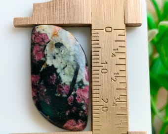 Puede incluir: Una piedra preciosa pulida con una mezcla de colores, incluyendo rojo oscuro, negro y blanco. La piedra se mide con un calibrador de madera, de unos 3 cm de largo.