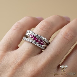 Puede incluir: Un anillo de plata con un diseño de doble banda. Una banda está engastada con pequeñas piedras preciosas rojas redondas, mientras que la otra está incrustada con pequeñas piedras preciosas claras. El anillo está en un dedo.