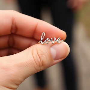 May include: A silver ring spelling out the word "love" in a delicate, cursive font. The ring is held between fingers, showcasing the elegant design. The ring's thin band and the word "love" are clearly visible.