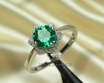 Puede incluir: Anillo de plata con una piedra verde brillante en el centro, rodeada por un engaste en forma de flor con pequeñas piedras claras. La fina banda pulida del anillo crea un aspecto clásico y elegante.