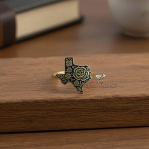 May include: A gold-toned ring featuring a dark blue Texas-shaped design with a floral rose and flower pattern. A smaller ring with a clear stone is also visible. The rings are displayed on a wooden surface.