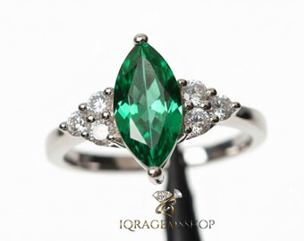 May include: A silver ring featuring a marquise-cut, vibrant green gemstone as the focal point, flanked by clusters of smaller, round, clear gemstones. The ring is set against a white background, highlighting the jewelry's sparkle and design. The text "IQRA GEMSSHOP" is visible.