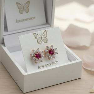 May include: A pair of flower-shaped earrings with a central red gemstone surrounded by clear crystals, presented in a white box. The earrings are gold-toned and set against a white card with the text "JORGEMSSHOP" and a butterfly illustration.