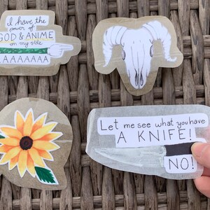 Puede incluir: Cuatro pegatinas en una superficie de mimbre marrón. Las pegatinas son: una calavera de carnero blanca, un girasol amarillo, una pegatina con el texto "Tengo el poder de DIOS & ANIME de mi lado AAAAAAAAA", y una pegatina con el texto "Déjame ver qué tienes UN CUCHILLO! ¡NO!"