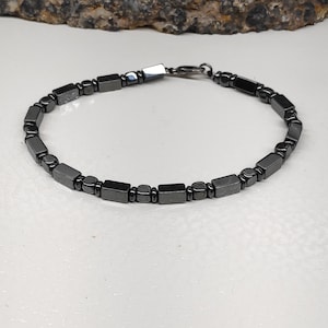 Puede incluir: Una pulsera gris oscuro con una mezcla de cuentas rectangulares y redondeadas. La pulsera está hecha de pequeñas cuentas pulidas unidas por un cierre plateado. La pulsera se muestra sobre una superficie blanca.