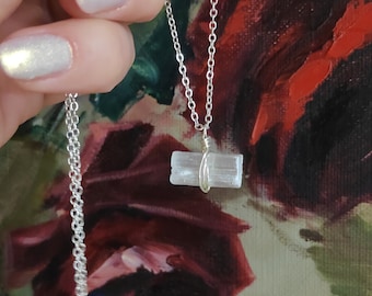 May include: A silver chain necklace with a clear selenite crystal pendant. The crystal is wrapped in silver wire and has a rectangular shape.