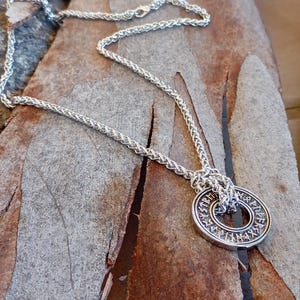 May include: A silver chain necklace with a silver pendant featuring a circle with runes inscribed around the edge. The pendant is a double circle with the chain running through the center.
