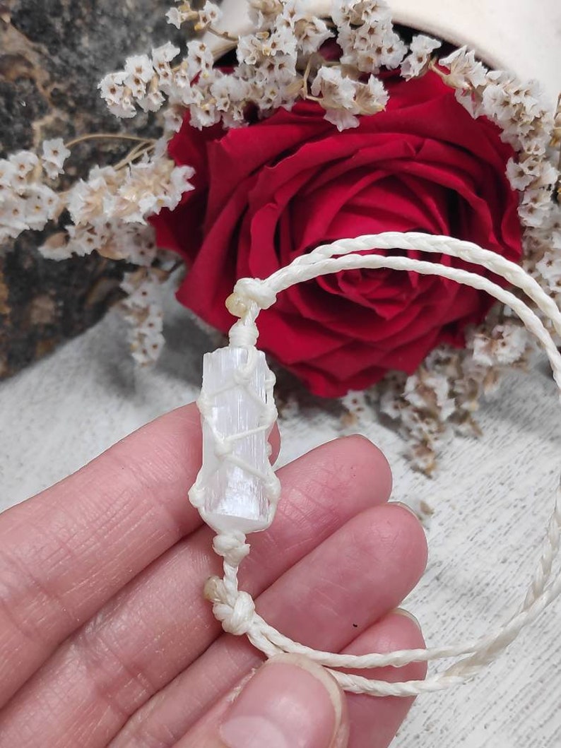 K&ouml;nnte beinhalten: Ein wei&szlig;es Selenit-Kristall, das in wei&szlig;es Makramee-Schnur gewickelt ist, bildet ein Armband. Das Armband ist mit einem Knoten gebunden und wird auf einer Hand gezeigt.