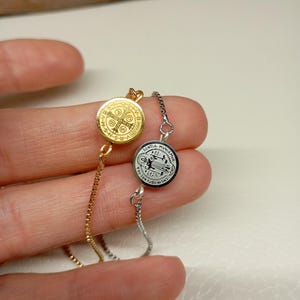 Puede incluir: Dos delicados brazaletes de cadena con medallones redondos. Un medallón es de oro con un diseño de cruz y el texto "S.P.D.  INRI" y el otro es de plata con una figura de mujer y el texto "S. MARIA  AUXILIO  DEI".