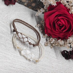 K&ouml;nnte beinhalten: Zwei Armb&auml;nder mit wei&szlig;en Kristallanh&auml;ngern, eines mit einem braunen Band und das andere mit einem wei&szlig;en Band. Die Armb&auml;nder liegen auf einer wei&szlig;en Oberfl&auml;che mit einer roten Rose und getrockneten Blumen im Hintergrund.