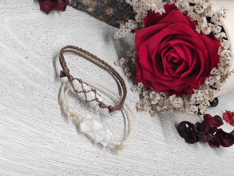 K&ouml;nnte beinhalten: Zwei Armb&auml;nder mit wei&szlig;en Kristallanh&auml;ngern, eines mit einem braunen Band und das andere mit einem wei&szlig;en Band. Die Armb&auml;nder liegen auf einer wei&szlig;en Oberfl&auml;che mit einer roten Rose und getrockneten Blumen im Hintergrund.