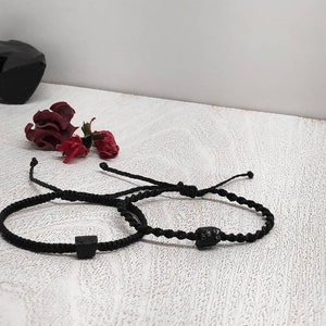 Könnte beinhalten: Zwei schwarze geflochtene Armbänder mit verstellbaren Bindungen. Eines hat eine quadratische schwarze Perle, das andere eine abgerundete schwarze Perle. Die Armbänder liegen auf einer weißen, strukturierten Oberfläche.
