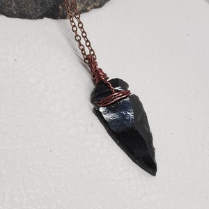 May include: A black obsidian arrowhead pendant wrapped in copper wire, hanging from a copper chain.